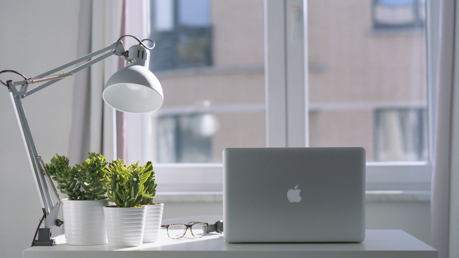 A minimalistic desk setup with a lamp, two potted plants, eyeglasses, and a laptop, in front of a window showing a building outside.
