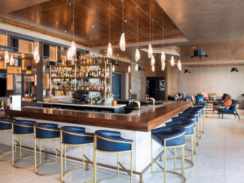 A modern bar with a large wooden counter, blue bar stools, hanging lights, and shelves filled with bottles, set in a stylish, open seating area.