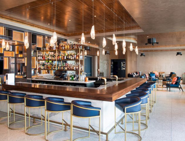 A modern bar setting with hanging lights, a well-stocked bar, blue bar stools with gold accents, and an inviting lounge area in the background.