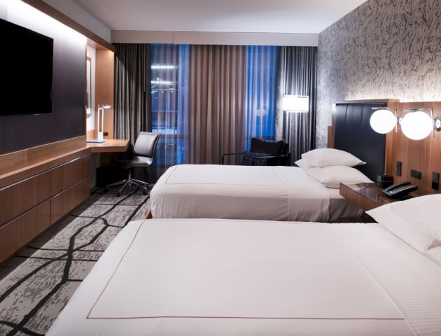 A modern hotel room with two beds, a large TV, a desk area, and stylish decor featuring patterned wallpaper and floor-to-ceiling curtains.