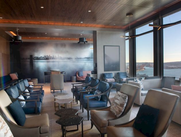 A modern lounge with comfortable seating, large windows displaying a water view, and a mural of a city skyline on the wall is shown.