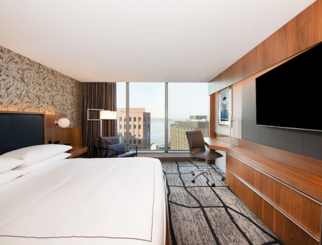 A modern hotel room with a large bed, a flat-screen TV, a desk, a chair, and floor-to-ceiling windows offering a city view, ending the sentence.