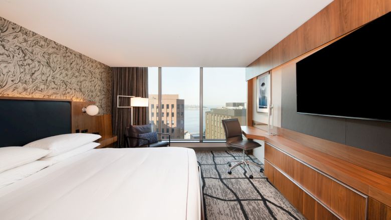 A modern hotel room with a large bed, flat-screen TV, desk, chair, and city view through large windows.